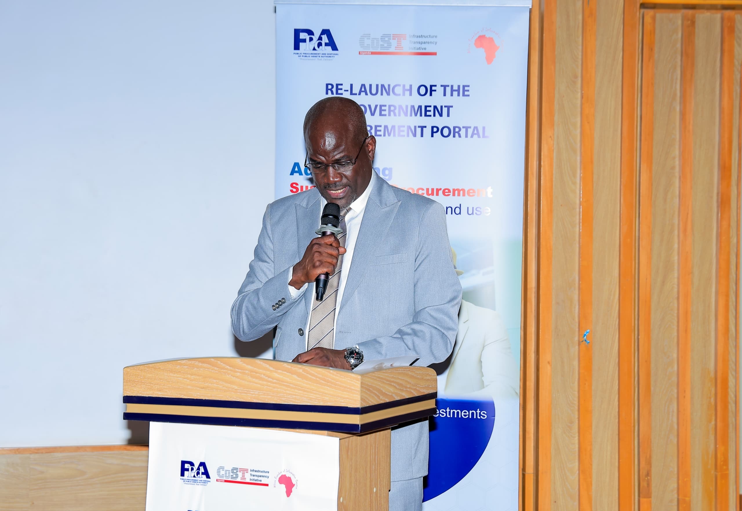A speaker addresses the relaunch of the Government Procurement Portal at the PPDA offices in Kampala, standing at a podium beside a banner reading RE-LAUNCH OF THE GOVERNMENT PROCUREMENT PORTAL.