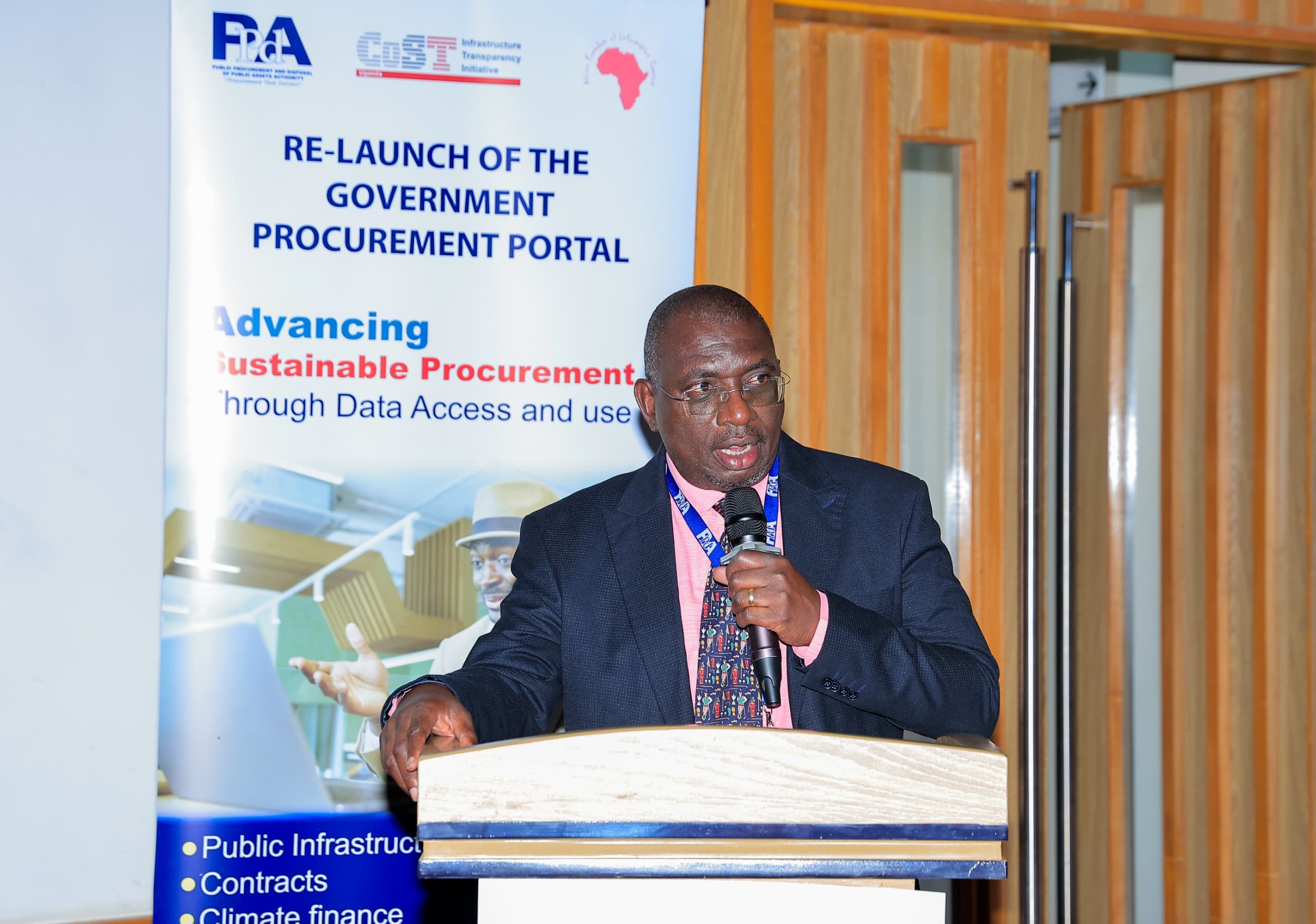A speaker at the podium beside a banner reading Advancing Sustainable Procurement Through Data Access and Use, listing Public Infrastructure, Contracts, and Climate Finance.